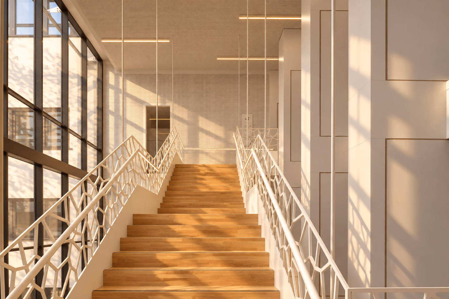 Le grand escalier du Site Botanique : des marches en chêne clair entre des garde-corps ajourés blancs, dans le soleil chaud de fin d’après-midi qui entre par de hautes fenêtres